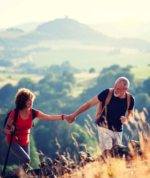 Senior tourist couple with backpacks hiking in nature, holding hands.
