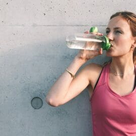A woman drinking CBD water against a concrete wall.